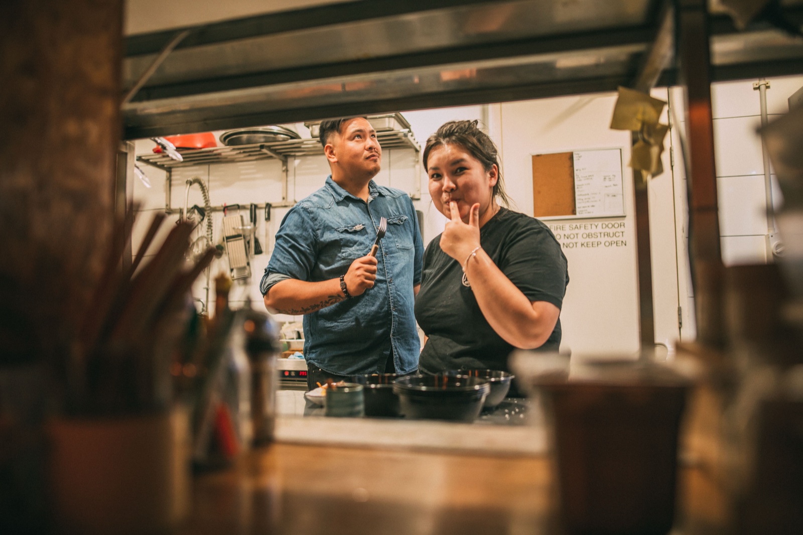 Andy Ngo and Rebecca Yii in the kitchen during service. Andy wears a denim shirt and holds tongs; Rebecca beside him in a black t-shirt.
